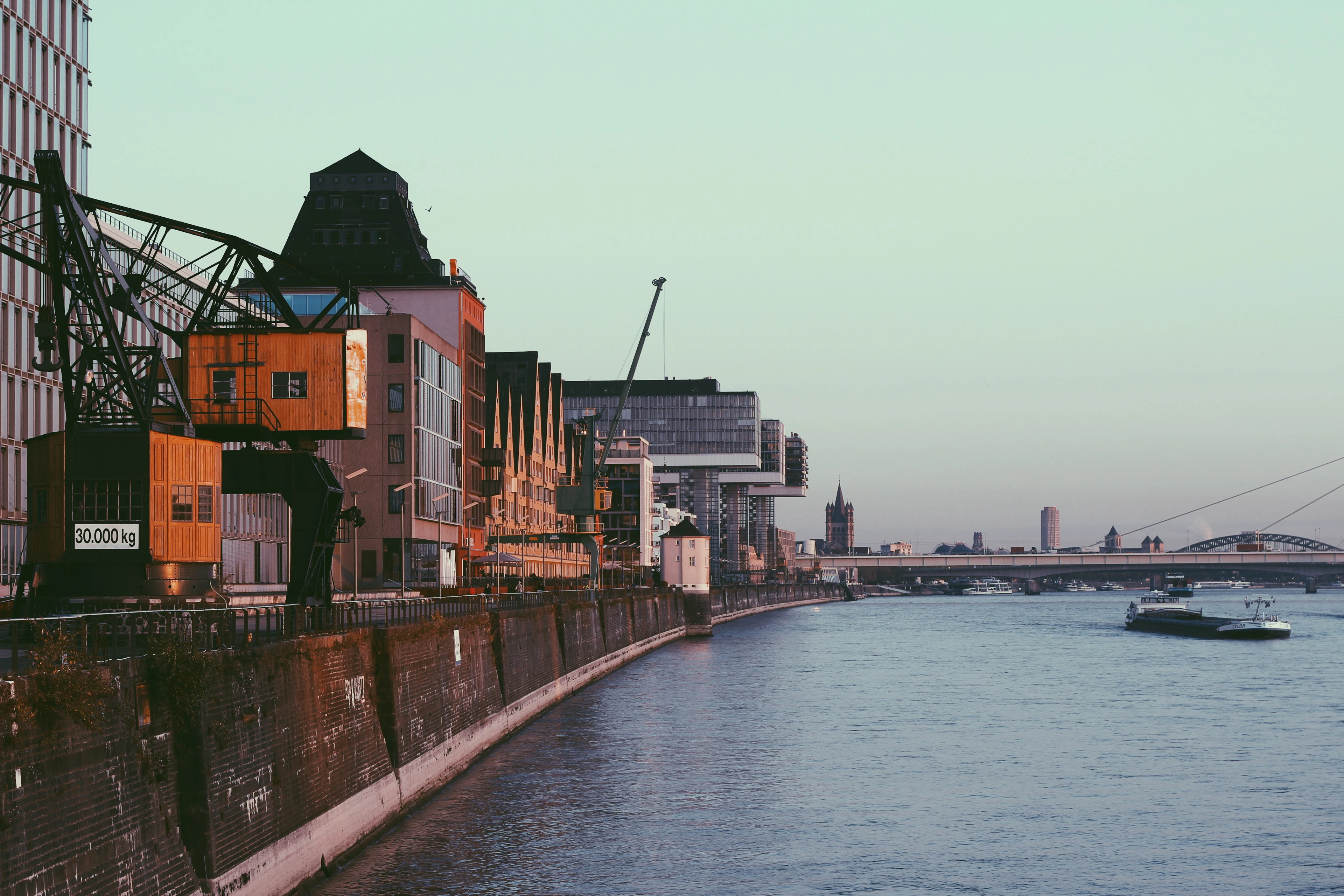 The Rhine at Cologne in morning light, historic black harbour cranes standing on the Rheinauhafen quay in the foreground and the glass crane-houses and a church tower ranged along the bank beyond, with a barge heading downstream