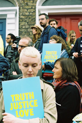 A group of people gathered outside with signs that read 'TRUTH JUSTICE LOVE' in bold yellow letters on a blue background. The crowd includes a diverse mix of individuals, some holding cameras, with expressions ranging from focused to contemplative. The background features red and blue doors with brick walls.