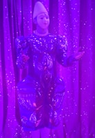 An individual wearing a decorative and elaborate costume stands against a backdrop of shimmering purple curtains. The outfit features intricate patterns with embellishments and is predominantly blue with detailed designs. A white headpiece complements their attire, and their expression suggests a performance setting.