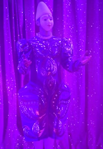 An individual wearing a decorative and elaborate costume stands against a backdrop of shimmering purple curtains. The outfit features intricate patterns with embellishments and is predominantly blue with detailed designs. A white headpiece complements their attire, and their expression suggests a performance setting.