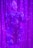 An individual wearing a decorative and elaborate costume stands against a backdrop of shimmering purple curtains. The outfit features intricate patterns with embellishments and is predominantly blue with detailed designs. A white headpiece complements their attire, and their expression suggests a performance setting.