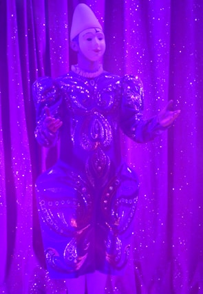 An individual wearing a decorative and elaborate costume stands against a backdrop of shimmering purple curtains. The outfit features intricate patterns with embellishments and is predominantly blue with detailed designs. A white headpiece complements their attire, and their expression suggests a performance setting.
