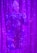 An individual wearing a decorative and elaborate costume stands against a backdrop of shimmering purple curtains. The outfit features intricate patterns with embellishments and is predominantly blue with detailed designs. A white headpiece complements their attire, and their expression suggests a performance setting.