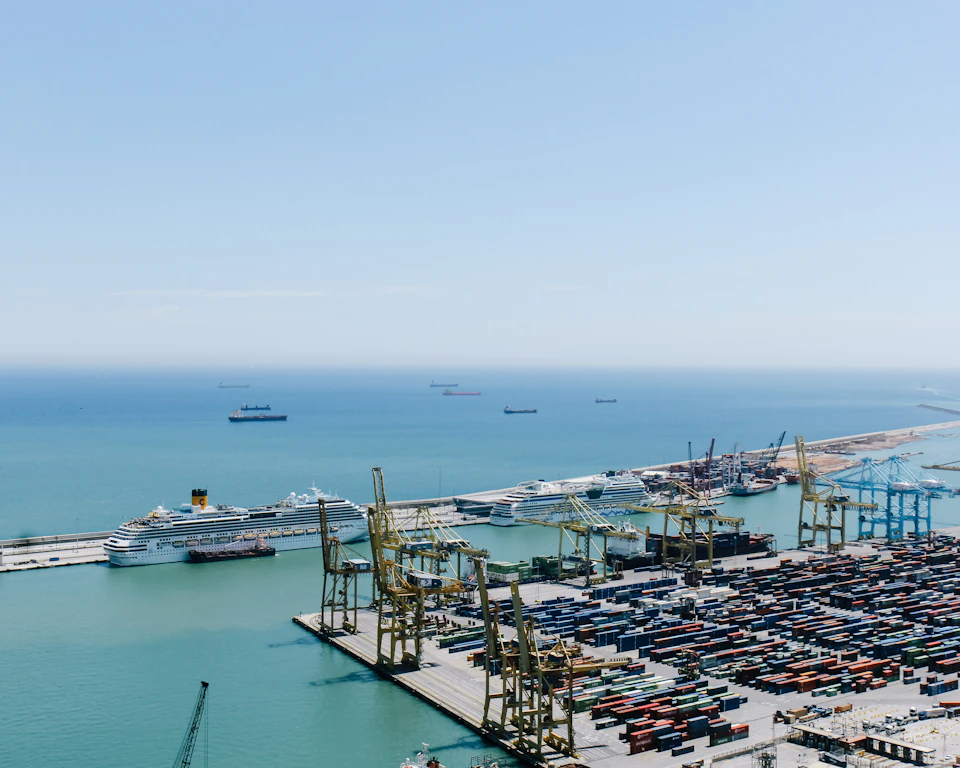 The image presents an aerial view of a bustling port, teeming with activity and filled with various types of ships and cranes. The port is situated on the coast, with the vast expanse of the ocean stretching out to meet it at the horizon. The sky above is a clear blue, providing a striking contrast to the greenish-blue hue of the water below. Numerous ships are docked in the harbor, their hulls painted in shades of white and gray, ready for loading or unloading cargo. The cranes, predominantly yellow and green, stand tall against this backdrop, their long arms reaching out towards the ships.
