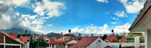 A panoramic view of a residential area with several houses featuring red-tiled roofs. The background displays a mountain range, while a bright blue sky with scattered clouds dominates the upper half of the scene.