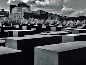A landscape with a series of large concrete rectangular slabs arranged in a grid pattern, set against an urban backdrop with modern buildings and a partly cloudy sky. Trees separate the slabs from the cityscape.