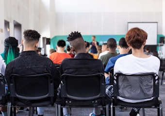 A group of people seated on chairs in what appears to be a conference or seminar setting. The individuals are facing forward, attentive to a presentation or speaker. The background features a projection screen and blurred figures of a person standing and speaking.