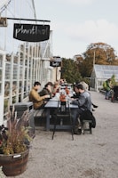 Several people are seated at a long outdoor table near a greenhouse, enjoying food and drinks. The table is lined with small potted plants, and there is a relaxed atmosphere as people engage in conversation. Nearby signage indicates the names of the establishments. The setting is surrounded by greenery and autumn foliage.