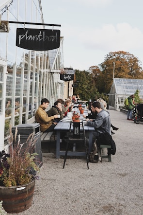 Several people are seated at a long outdoor table near a greenhouse, enjoying food and drinks. The table is lined with small potted plants, and there is a relaxed atmosphere as people engage in conversation. Nearby signage indicates the names of the establishments. The setting is surrounded by greenery and autumn foliage.
