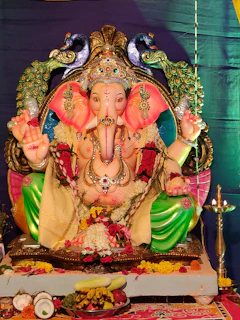 A colorful statue of Lord Ganesha adorned with intricate jewelry and vibrant clothing. The deity is seated, surrounded by peacocks, with offerings of fruits and flowers placed in front. The background displays a rich, deep purple curtain.