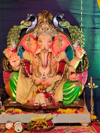 A colorful statue of Lord Ganesha adorned with intricate jewelry and vibrant clothing. The deity is seated, surrounded by peacocks, with offerings of fruits and flowers placed in front. The background displays a rich, deep purple curtain.