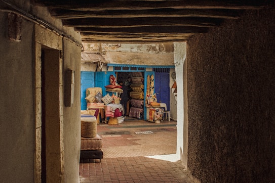 A narrow, dimly-lit alleyway leads to a vibrant market stall filled with colorful textiles and pillows. The stall is framed by bright blue doors, contrasting with the earthy tones of the alley. A variety of patterns and fabrics are displayed, inviting a sense of warmth and culture.