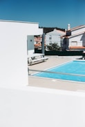 A modern outdoor scene featuring a bright blue swimming pool surrounded by beige tiles. In the foreground, white structures with clean lines contrast against the vivid sky. Residential buildings with red-tiled roofs are visible in the background, along with some greenery.