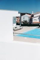 A modern outdoor scene featuring a bright blue swimming pool surrounded by beige tiles. In the foreground, white structures with clean lines contrast against the vivid sky. Residential buildings with red-tiled roofs are visible in the background, along with some greenery.