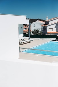 A modern outdoor scene featuring a bright blue swimming pool surrounded by beige tiles. In the foreground, white structures with clean lines contrast against the vivid sky. Residential buildings with red-tiled roofs are visible in the background, along with some greenery.