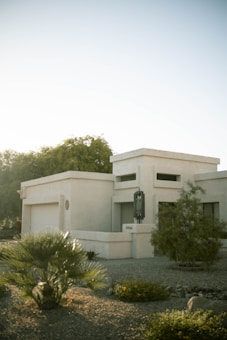 A modern, minimalist building features clean, geometric lines and a light-colored facade. There is sparse landscaping in the foreground with desert plants and gravel, suggesting a location in a dry climate. Soft sunlight casts gentle shadows on the structure, adding depth.