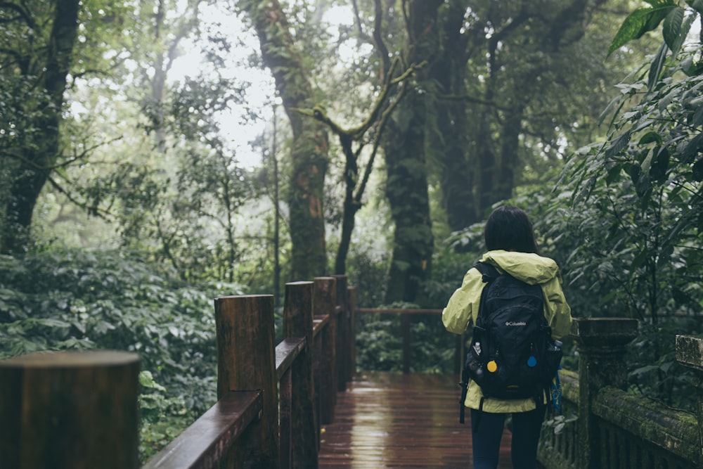 pessoa vestindo jaqueta verde com mochila em pé na ponte de madeira cercada de árvores altas e verdes durante o dia