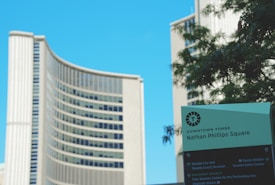 A modern, curved high-rise building with multiple floors and a glass facade is seen alongside a large green sign. The sign reads 'Downtown Yonge Nathan Phillips Square' and includes directions to nearby attractions such as Toronto City Hall and the Toronto Eaton Centre. There is a clear blue sky in the background, and some greenery with blurred leaves is visible near the sign.