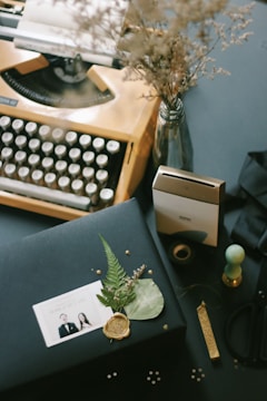 An eclectic desk arrangement featuring an old-fashioned typewriter with visible keys, a vase of dried flowers, a boxed gift adorned with a decorative wax seal and foliage, and an instant photo placed on top. Additional items include a rubber stamp, a tape dispenser, and other small stationary items scattered around.