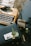An eclectic desk arrangement featuring an old-fashioned typewriter with visible keys, a vase of dried flowers, a boxed gift adorned with a decorative wax seal and foliage, and an instant photo placed on top. Additional items include a rubber stamp, a tape dispenser, and other small stationary items scattered around.