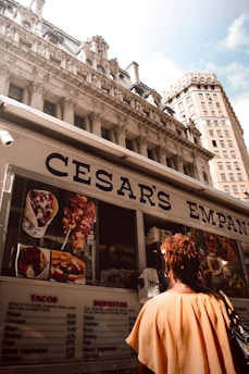 A food truck with a menu on the side offers items like tacos and burritos. The truck is positioned in front of a grand, historic building with intricate architectural details. A person wearing a light-colored top stands near the truck.