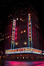 A brightly illuminated marquee reading 'Radio City Music Hall' displayed on a tall building at night. Neon lights in vibrant red, blue, and yellow colors enhance the facade, while streetlights reflect off a wet pavement. The building is decorated with vertical signs and flags, creating a lively nighttime atmosphere.