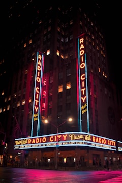 A brightly illuminated marquee reading 'Radio City Music Hall' displayed on a tall building at night. Neon lights in vibrant red, blue, and yellow colors enhance the facade, while streetlights reflect off a wet pavement. The building is decorated with vertical signs and flags, creating a lively nighttime atmosphere.