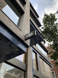 A modern building with multiple windows and a prominent sign that reads 'END.' The architecture features light brickwork and dark metallic frames, with reflective glass windows. A tree is visible on the right side, adding a touch of greenery. The perspective is angled upwards, capturing a section of the sky with clouds.