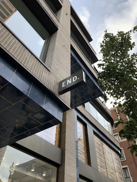 A modern building with multiple windows and a prominent sign that reads 'END.' The architecture features light brickwork and dark metallic frames, with reflective glass windows. A tree is visible on the right side, adding a touch of greenery. The perspective is angled upwards, capturing a section of the sky with clouds.