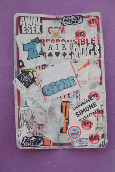 A collection of various stickers and graffiti are layered on a metal surface. The stickers have a mix of texts and images, with some partially torn or overlapping. The background is a vibrant purple wall.