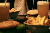 Two frothy beverages in tall glasses are placed alongside a burger with lettuce and tomato. A bowl of crispy fries is also visible, suggesting a casual dining scenario.