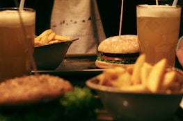 Two frothy beverages in tall glasses are placed alongside a burger with lettuce and tomato. A bowl of crispy fries is also visible, suggesting a casual dining scenario.