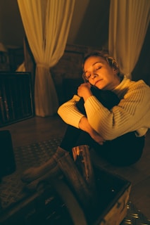 A person wearing a cozy sweater is sitting with eyes closed, embracing their knees in a warmly lit room with soft curtains and wooden accents.