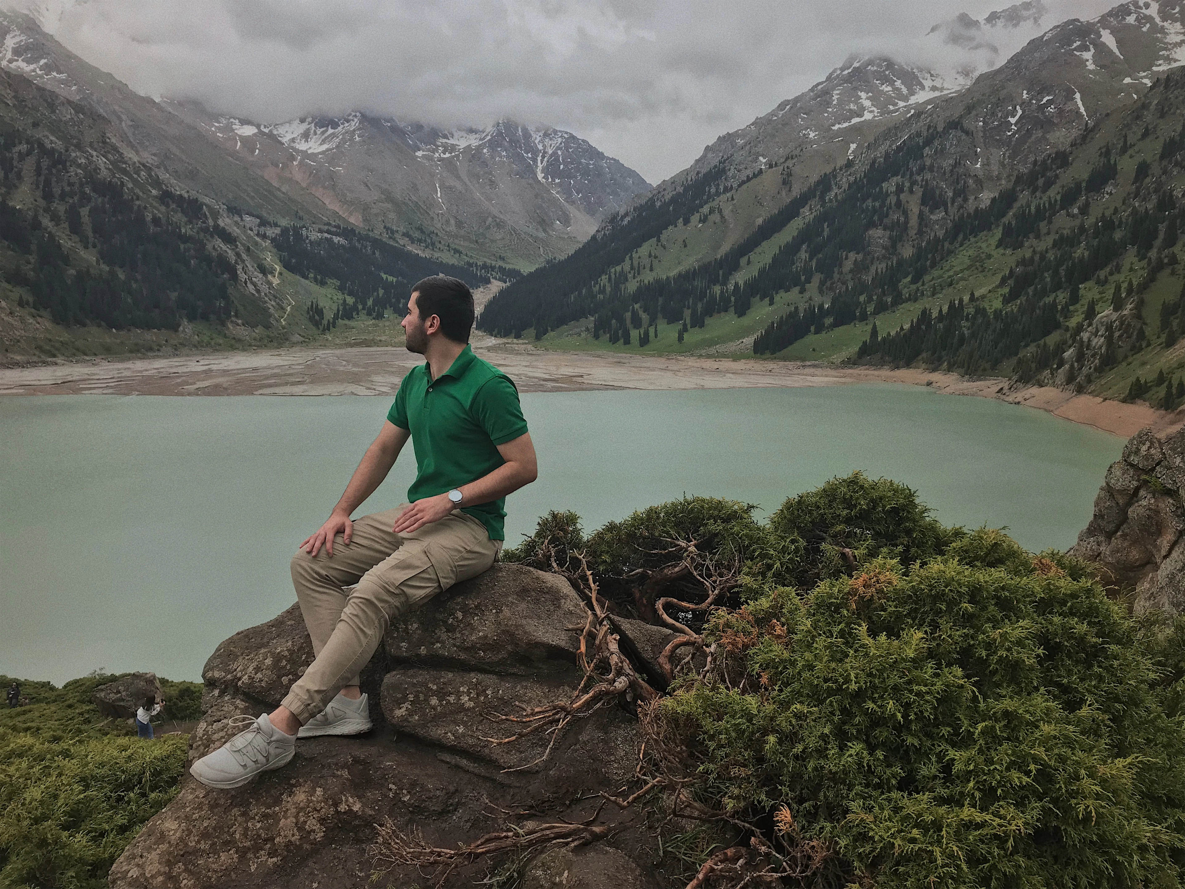 A hiker sits on a rock overlooking a turquoise alpine lake ringed by mountains near Almaty.