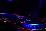 A dimly lit DJ setup with glowing buttons and dials, where hands are adjusting controls. The scene includes vibrant lights in blue and red hues on the equipment, suggesting an energetic atmosphere.