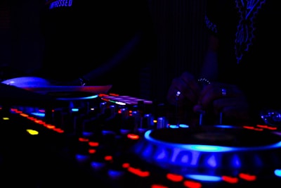 A dimly lit DJ setup with glowing buttons and dials, where hands are adjusting controls. The scene includes vibrant lights in blue and red hues on the equipment, suggesting an energetic atmosphere.