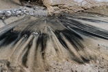 Sand mixed with dark streaks, possibly oil or ash, forms an intricate pattern alongside a stone surface on a beach. The area is surrounded by a combination of rough stones and smooth sand, creating an intriguing texture contrast.