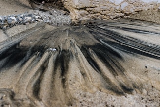 Sand mixed with dark streaks, possibly oil or ash, forms an intricate pattern alongside a stone surface on a beach. The area is surrounded by a combination of rough stones and smooth sand, creating an intriguing texture contrast.