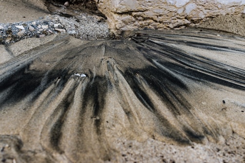 Sand mixed with dark streaks, possibly oil or ash, forms an intricate pattern alongside a stone surface on a beach. The area is surrounded by a combination of rough stones and smooth sand, creating an intriguing texture contrast.