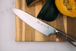 A sharp kitchen knife with a shiny blade and a metallic handle is placed on a wooden cutting board. Beside it, there are sliced and whole squash, showcasing their orange and green outer skins.