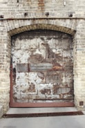 A large, weathered metal garage door set within a brick wall. The door shows significant signs of rust and deterioration, with patches of missing paint revealing bare metal underneath. The bricks are rough and textured, with some discoloration and rust stains particularly visible around the top of the doorway.