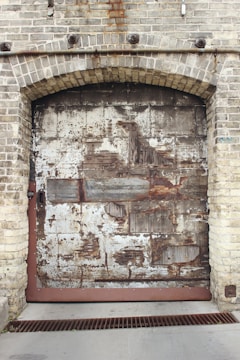 A large, weathered metal garage door set within a brick wall. The door shows significant signs of rust and deterioration, with patches of missing paint revealing bare metal underneath. The bricks are rough and textured, with some discoloration and rust stains particularly visible around the top of the doorway.