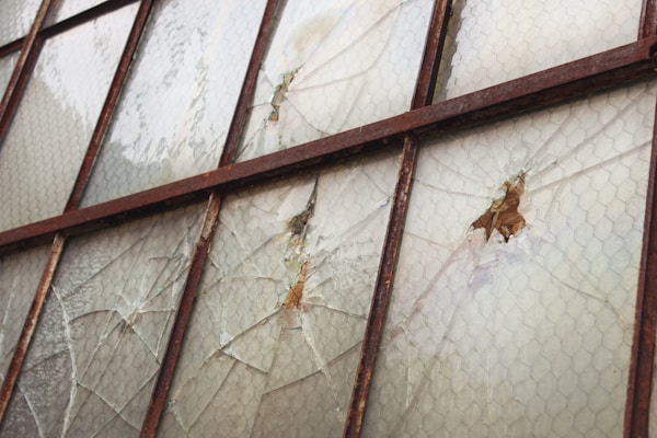 A series of weathered and cracked window panes, held together by a rusty metal framework. The glass has chicken wire embedded within, and some areas show significant damage with holes and missing fragments.
