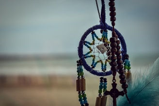 A dreamcatcher with a blue circular frame, adorned with colorful beads and a small pendant in the center. It is accompanied by wooden beads and a cross hanging alongside it. A blue feather is visible on the right side. The background is blurred, creating a soft, serene atmosphere.