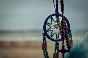 A dreamcatcher with a blue circular frame, adorned with colorful beads and a small pendant in the center. It is accompanied by wooden beads and a cross hanging alongside it. A blue feather is visible on the right side. The background is blurred, creating a soft, serene atmosphere.