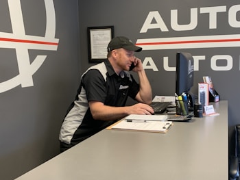 A man wearing a black and white uniform with a logo on the sleeve is sitting at a desk while talking on the phone. He is looking at a computer monitor in an office environment. There are various office supplies on the desk, including a pen holder, a stack of papers on a clipboard, and promotional materials.