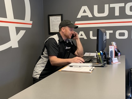 A man wearing a black and white uniform with a logo on the sleeve is sitting at a desk while talking on the phone. He is looking at a computer monitor in an office environment. There are various office supplies on the desk, including a pen holder, a stack of papers on a clipboard, and promotional materials.