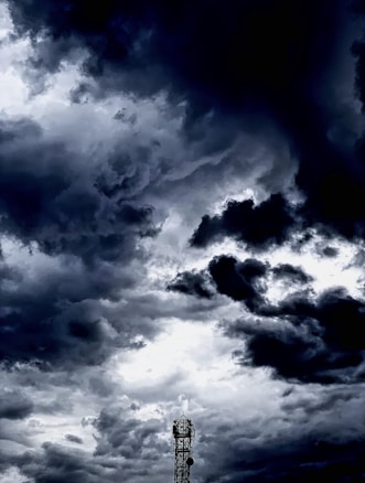 Dark, dramatic clouds fill the sky, creating a moody atmosphere. A solitary broadcast tower stands silhouetted against the swirling, stormy backdrop.