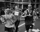 A group of runners participating in a city marathon or running event. They are captured mid-stride with expressions of focus and determination. The background features spectators and urban structures, indicative of a street race.