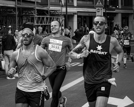 A group of runners participating in a city marathon or running event. They are captured mid-stride with expressions of focus and determination. The background features spectators and urban structures, indicative of a street race.